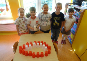 Pięcioro dzieci stoi przed stolikiem na którym ułożony jest uśmiech z białych i czerwonych kubeczków plastikowych.